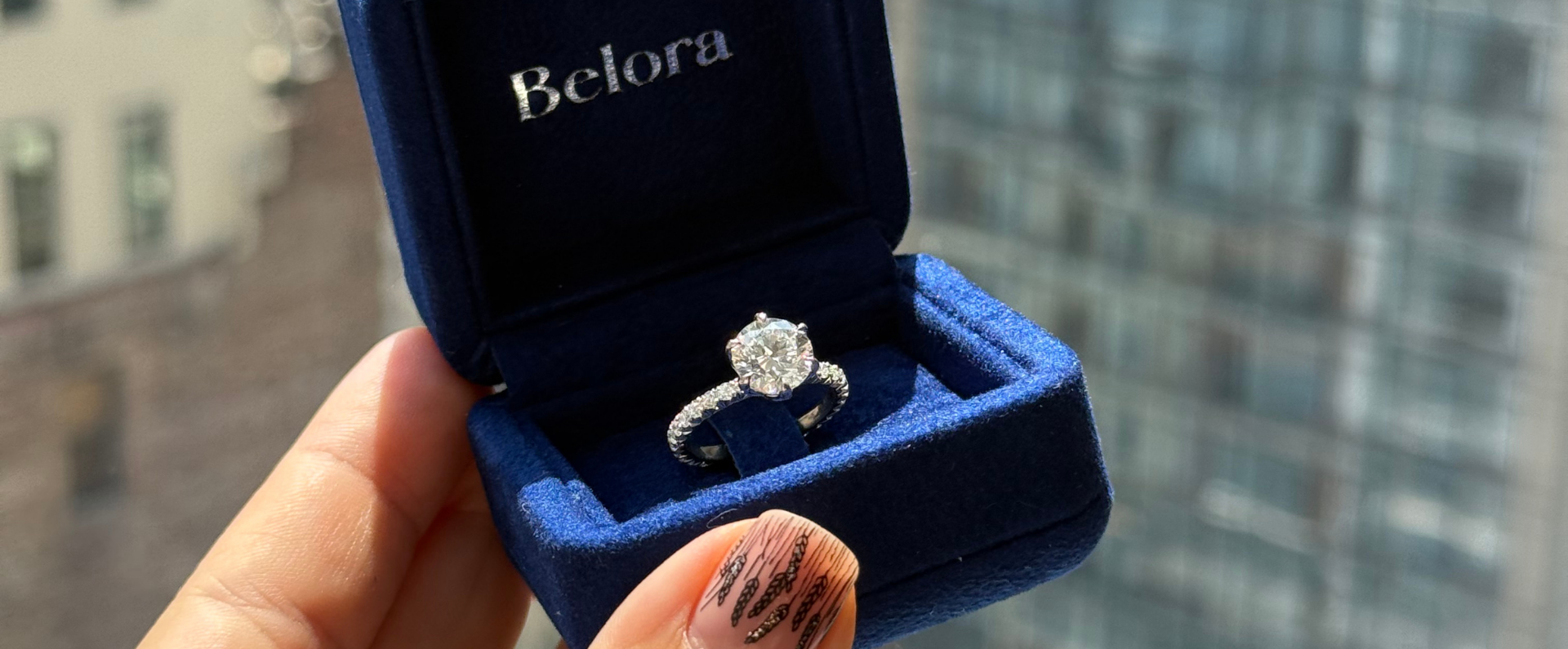 Belora Blue jewelry box with a ring inside, held by a hand with a blurred cityscape background