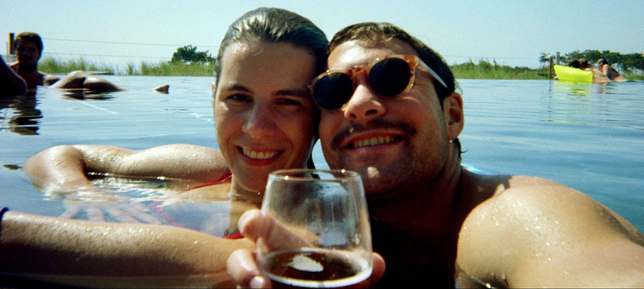 Belora Two people in a body of water, one holding a glass, with a clear sky background.